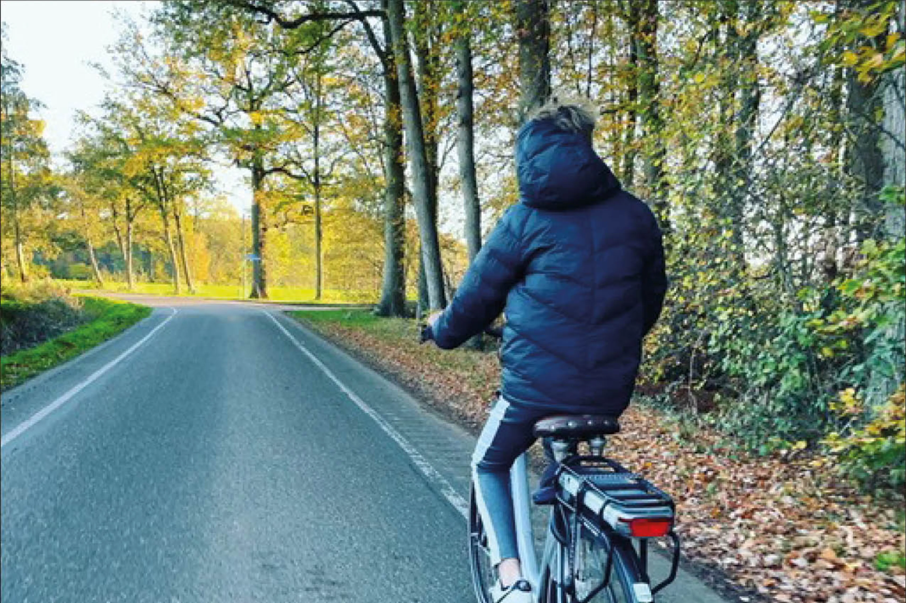 De zoon van Martine zit op zijn fiets en fietst op de weg. Hij draagt een dikke winterjas. Naast hem staan groene bomen en er liggen bruine bladeren in de berm. De zon schijnt in de verte.